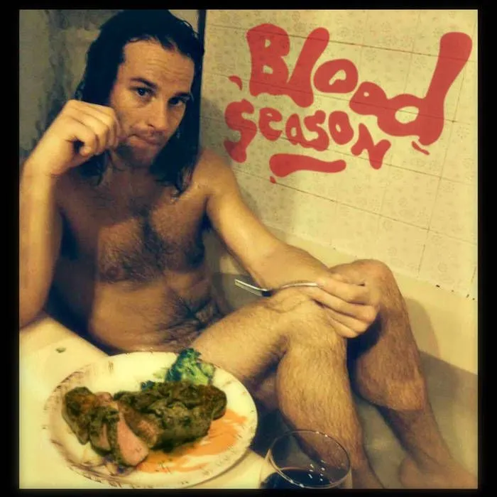 Lowell Thompson in a bathtub with a plate of food on his knee, the words Blood Season painted in red on the tiles behind him.