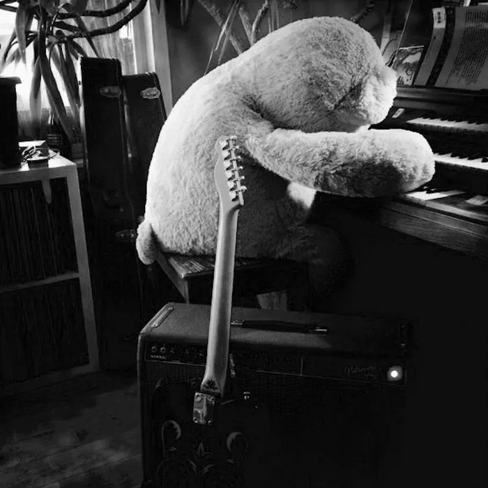 A black-and-white photograph of a teddy bear slumped over an upright piano, a guitar resting against an amplifier in front.