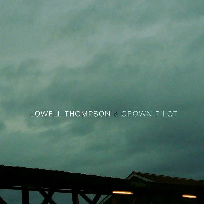 Storm clouds over a dark roofline with two lit windows, the artist name printed in white.