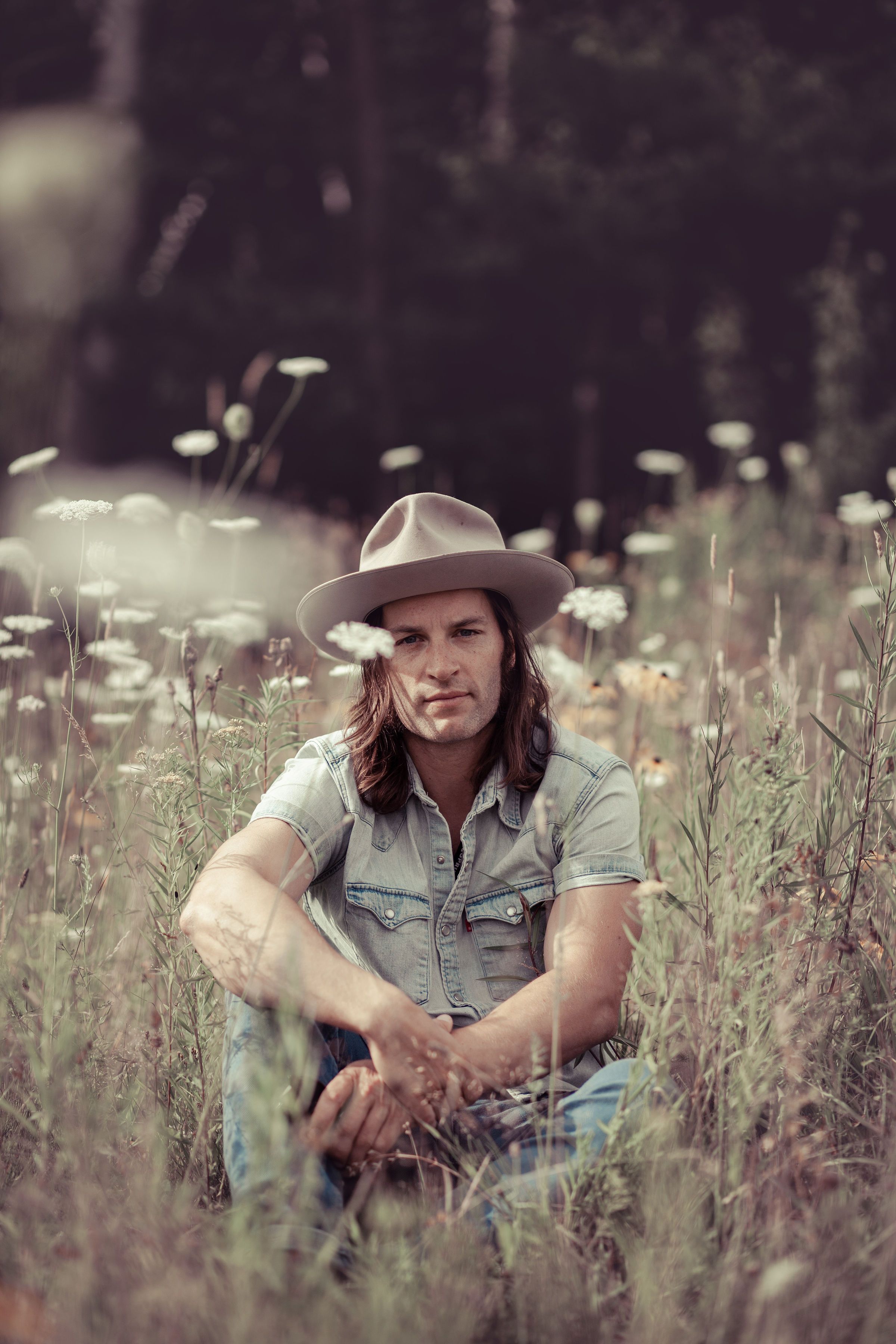Lowell Thompson sitting in a meadow of tall wildflowers, wearing a felt hat and a denim shirt, with dark forest behind him.